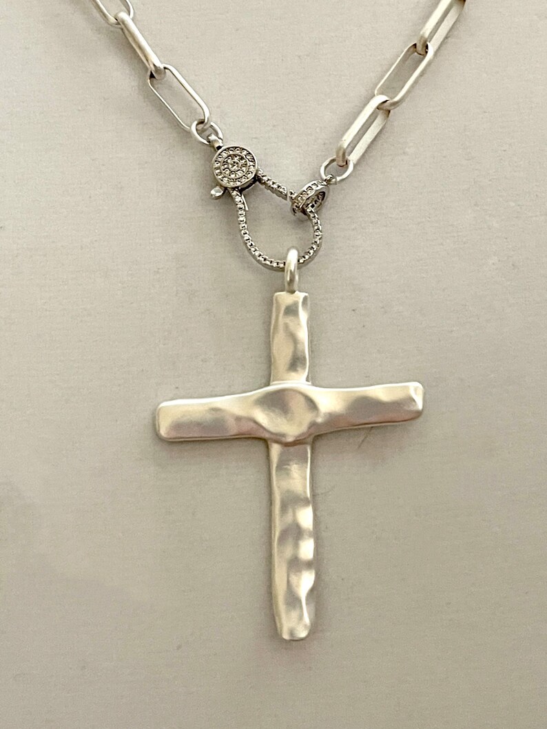May include: A silver chain necklace with a hammered silver cross pendant and a silver clasp with small clear stones.