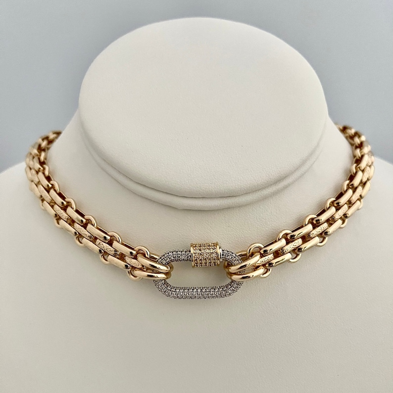 May include: A gold chain necklace with a large, oval-shaped clasp set with small diamonds. The chain is made of multiple strands of linked metal.