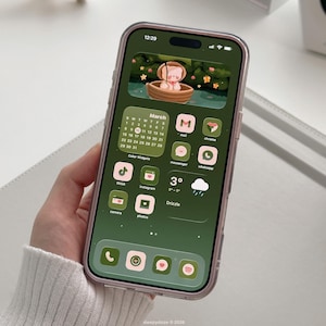 Puede incluir: Un smartphone con un tema verde y rosa, con una pantalla de inicio personalizada con iconos de aplicaciones y un widget de calendario de marzo. La pantalla muestra una ilustración de dibujos animados de un oso en un bote. El teléfono está en una funda transparente.