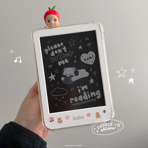 May include: A white Kobo e-reader in a clear case, decorated with small stickers of flowers and bears. The screen displays the text 'please don't bother me i'm reading' with a drawing of a bear reading, and a small apple-themed figure sits on top.