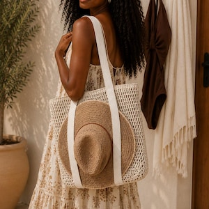 May include: A cream-colored woven tote bag with long white fabric straps, featuring a natural tan straw hat tucked into the front. The bag has an open-top design with a textured, grid-like pattern, providing a spacious interior for carrying items.