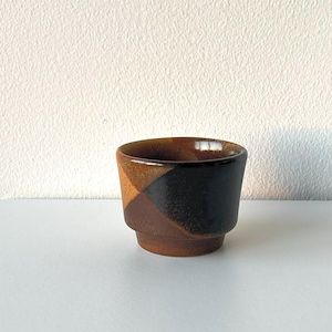 May include: A small, handcrafted ceramic cup with a geometric design. The cup features a combination of brown, black, and tan colors. The cup has a slightly flared rim and a stepped base, showcasing a minimalist aesthetic.
