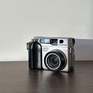May include: A silver Olympus Camedia C-2020 Zoom digital camera with a black lens and a black grip. The camera is sitting on a wooden surface.
