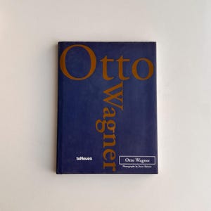 May include: A hardcover book titled "Otto Wagner" with a navy blue cover and gold lettering. The book's title is vertically oriented, with the author's name and publisher's logo at the bottom. The book is on a white surface.