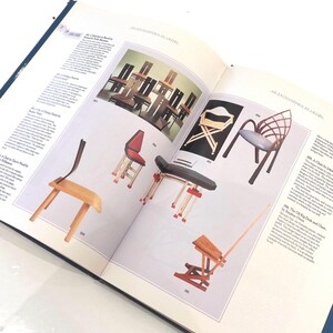 May include: Open book displaying various chair designs from "An Encyclopedia of Chairs." The pages showcase diverse chair styles, including a red-seated chair, a black and white folding chair, and a wooden chair with a long, thin arm.