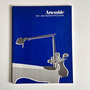 May include: A blue book with the title "Artemide 1999 - 2000 Master Price Book" and a black and white illustration of a lamp and a vase with flowers on a table.