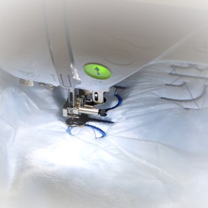 May include: A close-up of a white sewing machine embroidering a blue design on a white fabric. The machine has a green light indicating it is in use.