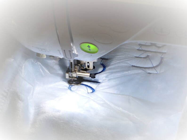 May include: A close-up of a white sewing machine embroidering a blue design on a white fabric. The machine has a green light indicating it is in use.