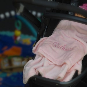May include: A pink baby blanket with the name "Ruchana" embroidered on it. The blanket is folded and placed in a black stroller.