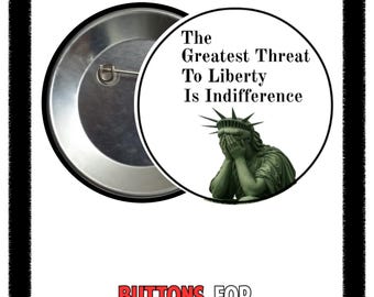 May include: A round button with a silver metal back and a white face. The face features the text "The Greatest Threat To Liberty Is Indifference" and an image of the Statue of Liberty. The words "Buttons For Democracy" are at the bottom.