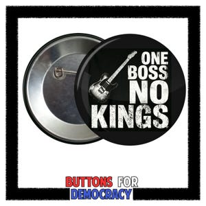 Op de afbeelding: Een zwarte knoop met de tekst "ONE BOSS NO KINGS" en een gitaarafbeelding. De knoop heeft een zilveren metalen achterkant en een speld. De woorden "BUTTONS FOR DEMOCRACY" staan onderaan.