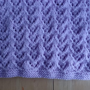 May include: A knitted lavender blanket with a detailed openwork pattern. The blanket features a repeating design of diamond shapes and a textured border. The soft, light purple color and intricate design suggest a handmade item, perfect for home decor or as a baby blanket.