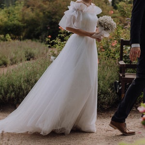 May include: A bride in a white tulle wedding dress with a ruffled neckline and a bouquet of white flowers. The groom is wearing a black suit and brown shoes. They are standing in a garden setting.
