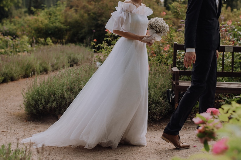 May include: A bride in a white tulle wedding dress with a ruffled neckline and a bouquet of white flowers. The groom is wearing a black suit and brown shoes. They are standing in a garden setting.