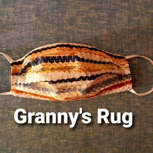以下が含まれることがあります： オレンジ、イエロー、ブラックのストライプのカラフルなパターンが施されたフェイスマスク。マスクはグラニー・スクエアのデザインで、マスクの下に「Granny's Rug」というテキストが印刷されています。