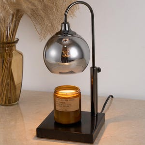 Puede incluir: Un calentador de velas de metal negro con una pantalla de vidrio plateado espejado. El calentador est&aacute; sobre una base negra y tiene una vela en &eacute;l. La vela es un tarro de vidrio marr&oacute;n con una etiqueta que dice "NESTLE GOLDEN COAST SOY CANDLE".