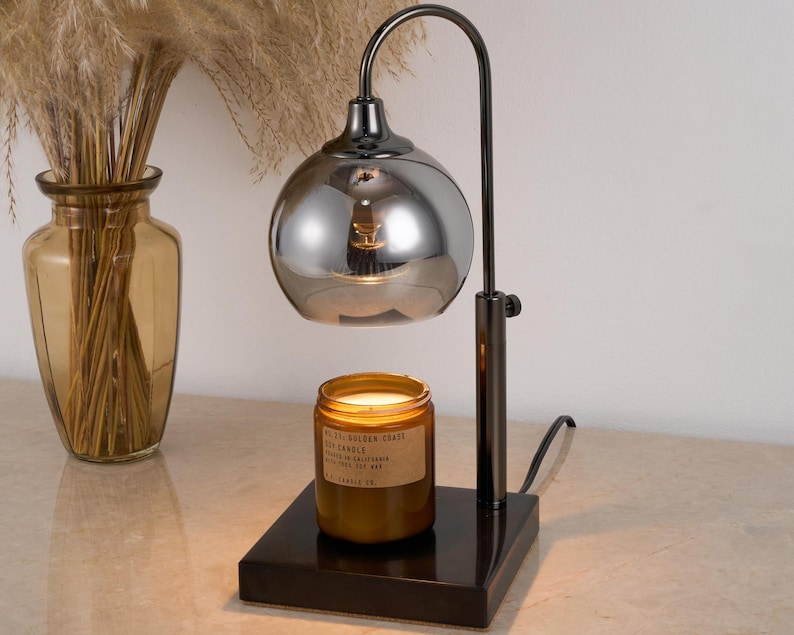 Puede incluir: Un calentador de velas de metal negro con una pantalla de vidrio plateado espejado. El calentador est&aacute; sobre una base negra y tiene una vela en &eacute;l. La vela es un tarro de vidrio marr&oacute;n con una etiqueta que dice "NESTLE GOLDEN COAST SOY CANDLE".