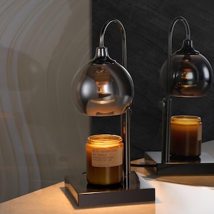 Puede incluir: Un calentador de velas de metal negro con una pantalla de vidrio y un tarro de vidrio marr&oacute;n con una vela. La vela est&aacute; etiquetada "VOLUSPA JAPANESE  CEREAL  SOY CANDLE  PEACH & NECTARINE  FRAGRANCE OIL  3.7 OZ (105G)".