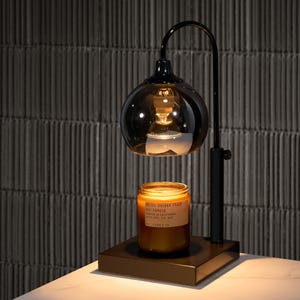 Puede incluir: Un calentador de velas de metal negro con una pantalla de vidrio y una bombilla. Un tarro de vela marr&oacute;n con el texto "MELT. OCEAN. FLOAT" est&aacute; sentado en el calentador.