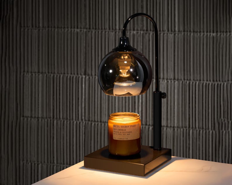 Puede incluir: Un calentador de velas de metal negro con una pantalla de vidrio y una bombilla. Un tarro de vela marr&oacute;n con el texto "MELT. OCEAN. FLOAT" est&aacute; sentado en el calentador.