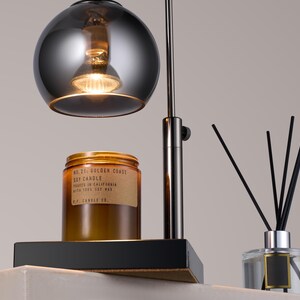 Puede incluir: Una l&aacute;mpara de mesa de metal negro con una pantalla redonda de vidrio plateado. Un tarro de vela de vidrio marr&oacute;n con el texto "No. 71 Golden Coast Soy Candle Poured in California With 100% Soy Wax P.F. Candle Co." est&aacute; sobre la mesa. Un difusor de vidrio transparente con ca&ntilde;as negras est&aacute; sobre la mesa.