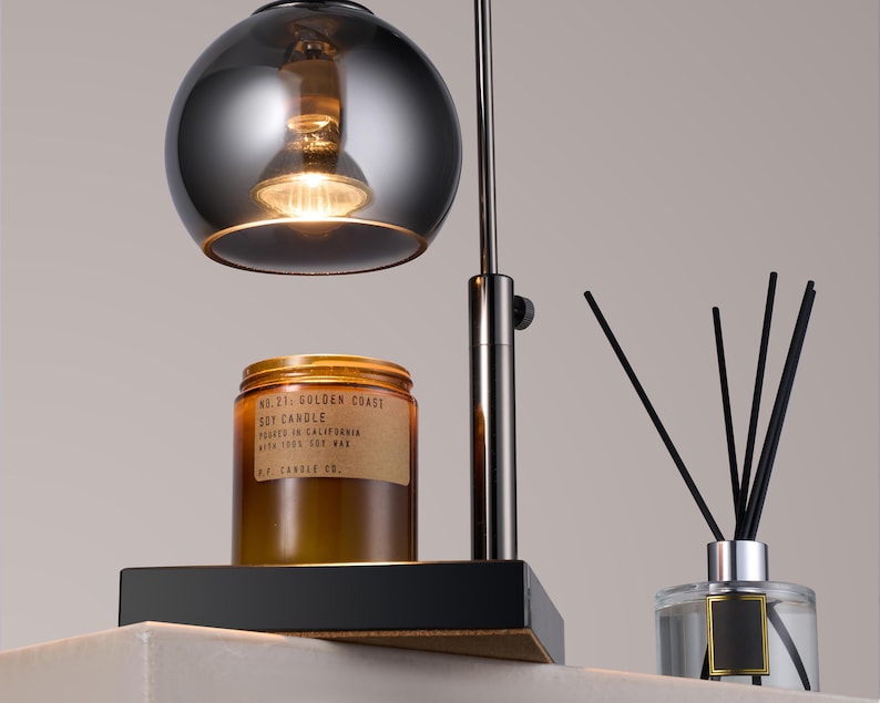 Puede incluir: Una l&aacute;mpara de mesa de metal negro con una pantalla redonda de vidrio plateado. Un tarro de vela de vidrio marr&oacute;n con el texto "No. 71 Golden Coast Soy Candle Poured in California With 100% Soy Wax P.F. Candle Co." est&aacute; sobre la mesa. Un difusor de vidrio transparente con ca&ntilde;as negras est&aacute; sobre la mesa.