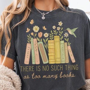 May include: Dark grey t-shirt with a whimsical design of books, flowers, and a hummingbird. The text "THERE IS NO SUCH THING as too many books" is printed below the books. The shirt is a comfortable, casual style.