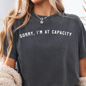 May include: A charcoal grey short-sleeved t-shirt featuring the white text SORRY, I'M AT CAPACITY printed in a clean, sans-serif font across the chest. The garment has a crew neckline and a slightly faded, vintage-inspired fabric finish.