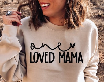 May include: A beige sweatshirt with the text "one loved mama" in black lettering. The text is in a cursive font with a heart symbol above the word "one".