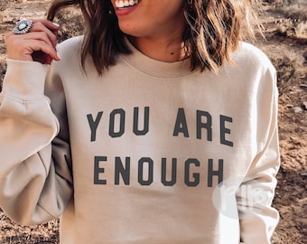Könnte beinhalten: Ein beiger Pullover mit dem Aufdruck "YOU ARE ENOUGH" in Schwarz.