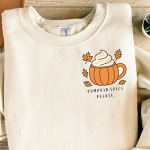 May include: Cream-colored sweatshirt with a graphic of a pumpkin spice latte. The design features an orange pumpkin-shaped mug with whipped cream and fall leaves, and the text "PUMPKIN SPICE PLEASE."