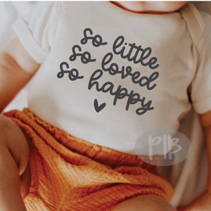 Puede incluir: Un body blanco para beb&eacute; con el texto "So little, so loved, so happy" en letra negra. Un peque&ntilde;o coraz&oacute;n est&aacute; debajo del texto. El body lo lleva un beb&eacute; con pantalones naranjas.
