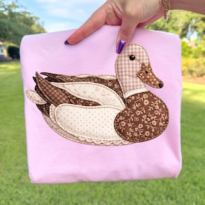 Op de afbeelding: Een lichtroze sweatshirt met een eendenapplicatie. Het eendenontwerp maakt gebruik van stoffen met patronen zoals ruitjes, bloemen en stippen in bruin, beige en crème. De eend is het centrale ontwerpelement op het sweatshirt.