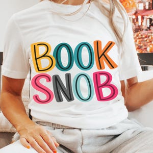 May include: White t-shirt with the text "BOOK SNOB" in colorful block letters. The text is in yellow, teal, orange, pink, and gray. The shirt is a casual style.
