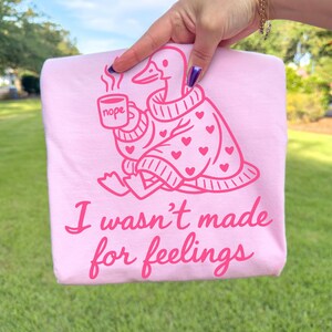 May include: A light pink t-shirt featuring a cartoon duck wearing a sweater with hearts, holding a mug that says "nope." The text "I wasn't made for feelings" is below the duck illustration.