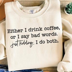May include: Cream-colored sweatshirt with the text "Either I drink coffee, or I say bad words. Just kidding. I do both." in black lettering. The sweatshirt is folded and displayed on a wooden surface.