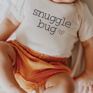 Puede incluir: Un body blanco para bebé con el texto "snuggle bug" y un pequeño corazón.