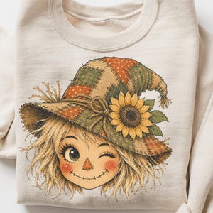 Può includere: Una felpa girocollo color crema con un'illustrazione grafica di un volto di spaventapasseri che fa l'occhiolino. Lo spaventapasseri indossa un cappello in stile patchwork nei toni dell'arancione e del verde, decorato con un grande girasole giallo.
