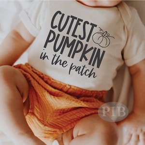 May include: A white baby onesie with the text "Cutest Pumpkin in the patch" and a small pumpkin graphic. The baby is wearing orange shorts.