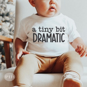 May include: A white baby onesie with the text "a tiny bit DRAMATIC" printed on the front. The baby is wearing brown pants and is sitting on a white chair.
