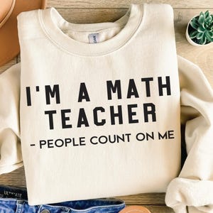 Op de afbeelding: Crèmekleurige sweatshirt met de tekst "I'M A MATH TEACHER - PEOPLE COUNT ON ME" in zwarte blokletters. De sweatshirt is opgevouwen, een deel van de mouw is zichtbaar. Een kleine vetplant in een witte pot staat op de achtergrond.