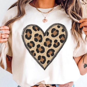 May include: Cream-colored t-shirt featuring a large heart design. The heart is filled with a leopard print pattern in shades of brown and black, outlined with a black border. The shirt has short sleeves and a crew neck.