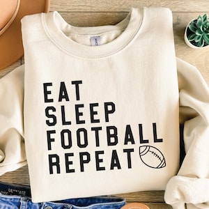 May include: A cream-colored sweatshirt with black text that reads "Eat Sleep Football Repeat" and a black and white football graphic.
