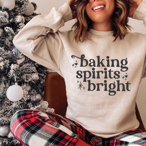 May include: A beige sweatshirt with the text "baking spirits bright" in black lettering with decorative elements. The sweatshirt is worn by a person with long brown hair and is paired with red and black plaid pants.