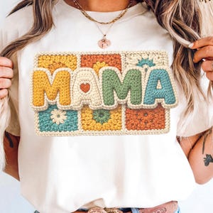 Op de afbeelding: Crèmekleurig T-shirt met het woord "MAMA" in een 3D-haakontwerp. De letters zijn geel, groen en blauw, met een hartdetail op de "A". Het ontwerp staat tegen een achtergrond van kleurrijke haakvierkanten.