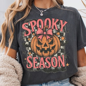 May include: Dark gray t-shirt featuring a distressed graphic with the words "SPOOKY SEASON" in pink, a jack-o'-lantern with a pink bow, and orange stars. The shirt has a vintage, worn look, perfect for Halloween.