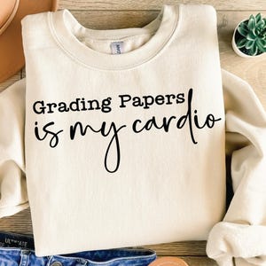 Może przedstawiać: Kremowa bluza z czarnym napisem "Grading Papers is my cardio". Bluza jest złożona, z małą rośliną sukulentową widoczną w tle. Odzież wykonana jest z miękkiego materiału.