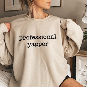 Könnte beinhalten: Ein beiger Pullover mit dem Aufdruck "professional yapper" in Schwarz.