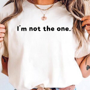 May include: Cream-colored t-shirt with the phrase "I'm not the one." printed in black. The shirt is a casual style, suitable for everyday wear. The image shows the upper body of a person wearing the shirt.