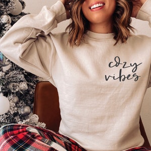 May include: A beige sweatshirt with the text "cozy vibes" written in black cursive on the front.